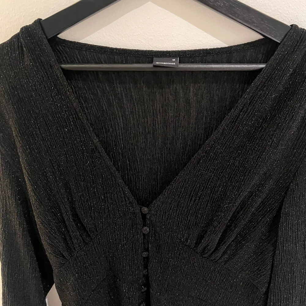 Snygg svart klänning från Gina tricot med glittrigt material och v-ringad design. Klänningen har långa ärmar och knappar framtill. Perfekt för en kväll ute eller en speciell tillställning.. Mekot.