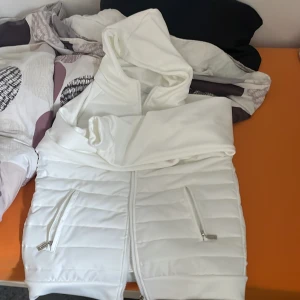 Vit quiltad väst med huva storlek 165/170 - Snygg vit quiltad väst med dragkedja och huva. Perfekt för kyliga dagar när du vill hålla stilen. Västen har praktiska fickor med dragkedjor för säker förvaring.
