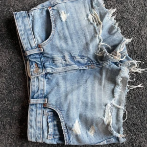 Blå jeansshorts från Pull&Bear - Snygga blå jeansshorts från Pull&Bear med slitna detaljer och fransiga kanter. Perfekta för en avslappnad stil. De har en klassisk femficksdesign och knappgylf. Använda en gång, säljer då dem är för små