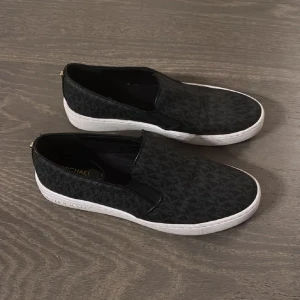 Svarta slip-on skor från Michael Kors - Snygga svarta slip-on skor från Michael Kors med ett diskret mönster och vit sula. Perfekta för en stilren look. Skorna har en bekväm passform och är lätta att ta på och av. Köpt sacond hand ingen låda därav priset