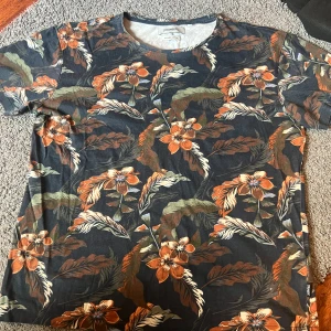 Blommig t-shirt från Strömberg - Snygg t-shirt från Strömberg i ekologisk bomull med ett färgglatt blommönster. Perfekt för att liva upp garderoben med sina orange och gröna toner. Den har en rund hals och korta ärmar.