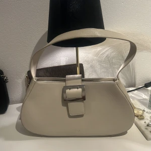 Beige axelväska med spänne - Snygg beige axelväska i skinn med ett stilrent spänne framtill. Väskan har en praktisk dragkedja på baksidan och flera fack inuti för enkel organisering. Perfekt för att bära med sig det viktigaste med stil.