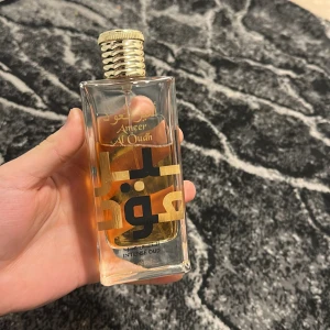Ameer Al Oudh Intense Oud Parfym - Säljer en elegant parfymflaska med Ameer Al Oudh Intense Oud. Flaskan är rektangulär med guldlock och har en stilren design med arabiska bokstäver i svart och guld. Perfekt för den som älskar djupa och intensiva dofter.
