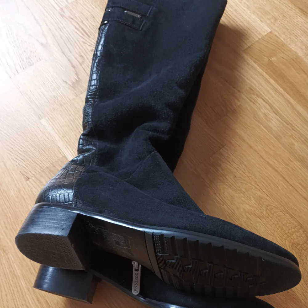 Snygga svarta stövlar i mocka med krokodilmönstrade detaljer och dragkedja på sidan.vinterstövlar i äkta läder med äkta päls inuti. Perfekta för en stilren look. De har en låg klack och är knähöga, vilket ger en elegant touch. . Kengät.