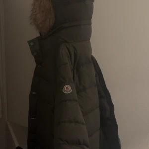 Olivgrön dunjacka från Moncler - Säljer en olivgrön dunjacka från Moncler med pälskantad huva. Jackan har en quiltad design och stängs med både dragkedja och knappar. Den har praktiska fickor framtill och en broderad logotyp på ärmen. Perfekt för kalla dagar!
