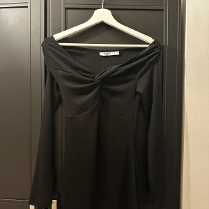 Svart offshoulder klänning från NA-KD - Snygg svart offshoulder klänning från NA-KD i ribbat material. Perfekt för festliga tillfällen med sin eleganta design och långa ärmar. Klänningen har en knutdetalj framtill som ger en extra touch. Passar perfekt för vår och sommar.