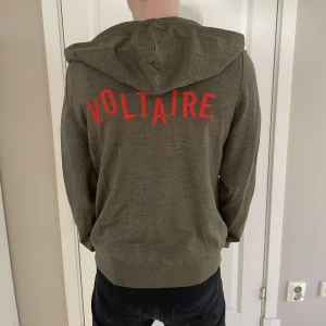 Zadig & Voltaire ziphoodie - Säljer en snygg olivgrön ziphoodie av merionoull från Zadig & Voltaire. Den har en dragkedja framtill och texten 'VOLTAIRE' i rött på ryggen. Perfekt för en casual look och passar bra till både höst och vår. Den är i bra skick och superbekväm att ha på sig. Perfekt för dig som gillar stil och komfort! 🌟Storlek S men passar M!
