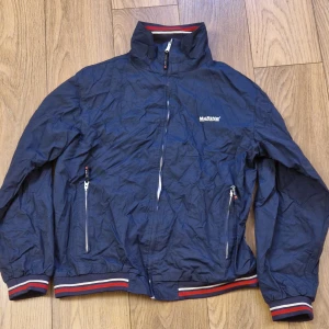 Marinblå jacka från Marine - Säljer en marinblå jacka från Marine Scandinavian Sportswear i storlek XL. Jackan har en dragkedja framtill och två fickor med dragkedjor. Den har ribbade muddar med röd och vit randdetalj vid ärmslut och nederkant.
