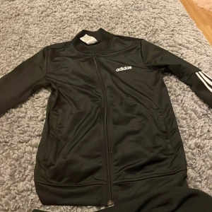 Svart träningsjacka från Adidas - Snygg svart träningsjacka från Adidas med klassiska vita ränder längs ärmarna. Jackan har en dragkedja framtill och är tillverkad i ett syntetmaterial som ger en sportig look. Perfekt för träning eller en avslappnad stil.