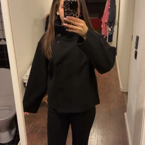 Svart kappa med knappar - Säljer en stilren svart kappa i mycket bra skick. Den har en hög krage och knappar framtill. Perfekt för höst och vinter, och ger en elegant look. Passar bra till både vardag och fest. Storleken är normal och den har långa ärmar.