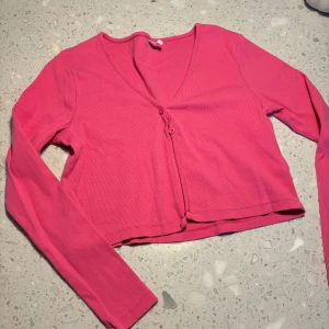 Rosa ribbad kofta från ONLY - Säljer en snygg rosa ribbad kofta från ONLY. Den har en v-ringning och knappar framtill. Perfekt för att lägga till en färgklick i din garderob! Sitter som en M