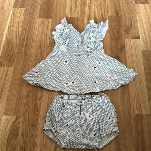 Vit och blå babyklänning med volanger från Ebbe - Söt vit och blå babyklänning från Ebbe med små mönster och volangdetaljer. Klänningen har en luftig passform och matchande byxor med resår i midjan. Perfekt för varma dagar!