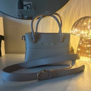  Valentino väska - Elegant beige handväska från Valentino med avtagbar axelrem och dekorativt metallspänne. Väskan har två handtag och en dragkedja för säker förvaring. Perfekt för en stilren look. Nypris 2000 kr