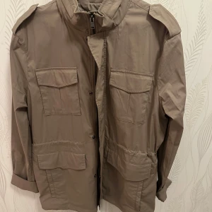 Beige jacka med dragkedja - Säljer en beige jacka med dragkedja och knappar. Jackan har långa ärmar och flera praktiska fickor framtill. Perfekt för höst och vår. Inbyggd luva