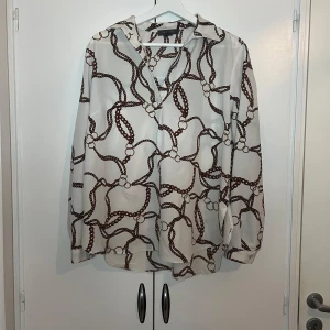 Vit och brun mönstrad blus - Snygg vit blus med brunt kedjemönster. Blusen har långa ärmar och en klassisk krage. Perfekt för en stilren look.