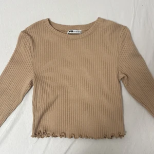 Beige ribbad tröja från FB Sister - Säljer en superfin beige ribbad tröja från FB Sister i storlek M. Den är croppad med långa ärmar och har en söt volangdetalj vid nederkanten. Perfekt för både höst och vår! 🧡