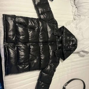 Svart pufferjacka med huva - Snygg svart pufferjacka från Moncler med glansig finish och praktisk huva. Jackan har en dragkedja framtill och är perfekt för kyligare väder. Den quiltade designen ger extra värme och stil. storlek xl men den är liten i storlek 