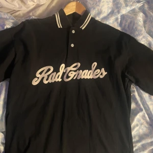 Svart pikétröja från H&M - Säljer en svart pikétröja från H&M med texten 'Rad Grades' i vitt på framsidan. Tröjan har en oversized passform och klassisk krage med vita ränder. Perfekt för en avslappnad stil!