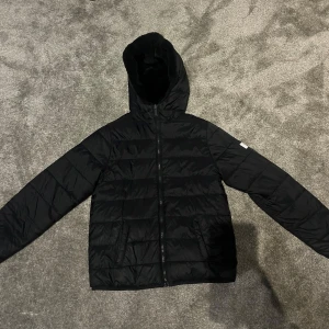 Svart pufferjacka med huva - Säljer en svart pufferjacka i mycket bra skick. Den har en praktisk huva och dragkedja framtill. Perfekt för kalla höst- och vinterdagar. Jackan är långärmad och har en stilren design som passar till det mesta. Skön och varm, idealisk för både vardag och utflykter. Kan has åt båda hål så de blir antingen dun eller pälsjacka. Barnstorlek, vet dock inte vilken, passar XS eller 12-14 år. Litet hål på ena armen de sista bild 