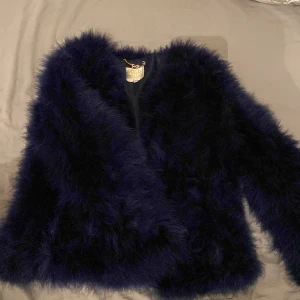 Mörkblå pälsjacka från Pello Bello - Säljer en supermjuk och fluffig mörkblå pälsjacka från Pello Bello. Perfekt för att hålla sig varm och stilren under kyliga kvällar. Jackan har en elegant design utan knappar eller dragkedja, vilket ger en öppen och avslappnad look. Passar perfekt för både fest och vardag!