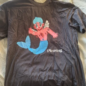 Svart t-shirt med sjöjungfrumotiv från Pleasing - Cool svart t-shirt från Pleasing med ett färgglatt sjöjungfrumotiv i rosa och blått. Storlek medium. Lite skrynklig pga legat i garderoben men fixar innan den skickas! Knappt använd. Köptes för 1000kr på pleasings pop up store i London. 