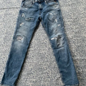 Blå jeans med slitningar från Replay - Säljer ett par snygga blå jeans från Replay med coola slitningar på framsidan. De har en klassisk femficksdesign och en läderdetalj på baksidan. Perfekta för en avslappnad stil! 🕶️