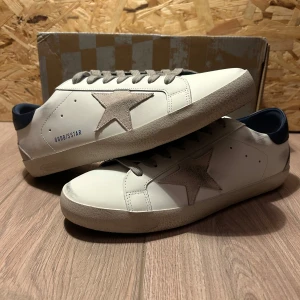 Golden Goose Sneakers - Snygga vita sneakers från Golden Goose med grå stjärndetalj på sidan och grå snörning. Skorna har en mörkblå häl och en klassisk design som passar perfekt för en stilren look. True to size. Hör av dig vid frågor- Emil 🤝