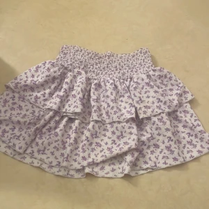 Ljuslila blommig kjol med volanger - Charmig ljuslila kortkjol med blommigt mönster och volanger. Perfekt för en somrig look med sin elastiska midja som ger en bekväm passform. Kan även använda som en top. 