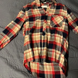 Rutig skjorta från These Glory Days - Snygg rutig skjorta i rött, beige och blått från These Glory Days. Skjortan har långa ärmar och en klassisk krage med knappar framtill. Perfekt för en avslappnad stil med en bröstficka som extra detalj.