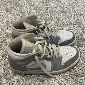 Nike Air Jordan 1 - Säljer ett par grå och vita Nike sneakers i bra skick. De har snörning och en klassisk design med perforerade detaljer på tån för extra ventilation. Perfekta för vardagsbruk eller en avslappnad stil. Skorna har lite slitage men är fortfarande snygga och bekväma.