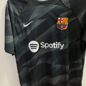  FC Barcelona fotbollströja - Säljer en svart FC Barcelona fotbollströja i storlek L från Nike. Tröjan har korta ärmar. Den har klubbens emblem och sponsorer som Spotify och Beko tryckta på framsidan. Perfekt för alla Barça-fans! Aldrig använd splitter ny