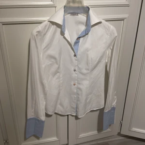 Vit skjorta med blå detaljer från Fagott - Elegant vit skjorta från Fagott med blå randiga detaljer på krage och ärmslut. Tillverkad i 100% bomull för en bekväm känsla. Perfekt för en stilren look.