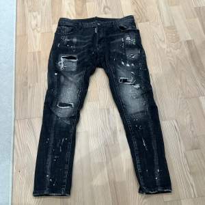 Säljer ett par svarta jeans från Dsquared2 i bra skick. De har en cool distressed look med slitningar och färgstänk. Jeansen har knappar i gylfen och en normal passform. Perfekta för en avslappnad och trendig stil!