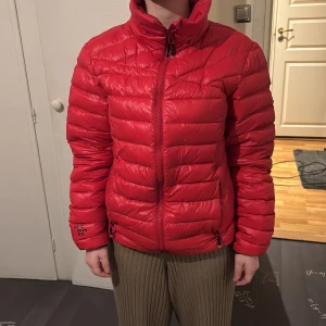 Röd pufferjacka från Neomondo - Snygg röd pufferjacka moncler liknande  från Neomondo med dragkedja och hög krage. Perfekt för kyliga dagar med sin vadderade design. Jackan har en normal passform och är långärmad.