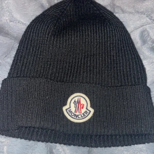Svart stickad mössa från Moncler - Snygg svart stickad mössa från Moncler med broderad logotyp framtill. Perfekt för kyliga dagar och ger en stilren look. Passar både till vardags och mer uppklädda tillfällen. 