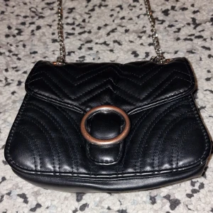 Svart quiltad axelväska med kedja - Snygg svart axelväska i quiltat mönster med en cool kedja som axelrem. Väskan har en rund metallring på framsidan som detalj och stängs med en tryckknapp. Perfekt för en utekväll eller när du vill ha en stilren look. 🖤