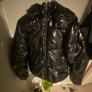 Svart glansig pufferjacka från Nelly - Säljer en svart glansig pufferjacka från Nelly. Den har en dragkedja framtill och en mysig huva. Perfekt för kalla dagar när du vill hålla dig varm och stilren. Jackan är i bra skick och redo för nya äventyr!