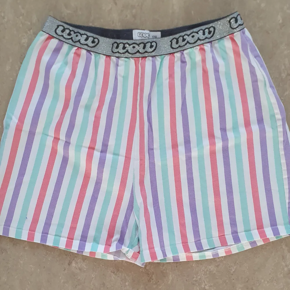 Färgglada randiga shorts från Wow i storlek 170. De har en elastisk midja med ett glittrigt band och texten 'wow'. Perfekta för sommarens alla äventyr med sitt mjuka och bekväma material.. Shorts.