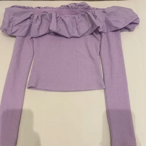 Lila off-shoulder topp med volang - Säljer en superfin lila off-shoulder topp med volangdetaljer. Toppen är ribbad och har långa ärmar. Perfekt för både vardag och fest! 💜