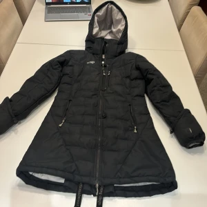 Svart dunjacka från Uhip - Säljer en svart, fodrad stalljacka från Uhip i storlek 150 barn. Jackan har en lång och figursydd design med en dragkedja framtill och två dragkedjefickor på sidorna. Den har en huva som går att ta bort och passar bra för kalla vinterdagar i stallet. Jackan är i bra skick och väldigt varm och bekväm. Perfekt för vintern! ❄️