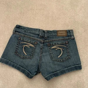 Jeansshorts från Frankie B. - Säljer ett par snygga jeansshorts från Frankie B. De är mörkblå och har en låg midja. Shortsen har två bakfickor med coola broderade detaljer och en knapp samt dragkedja framtill. Perfekta för sommaren! 🩳💙