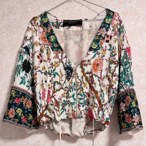 Blommig blus från Zara - Säljer en superfin somrig blus med blommönster från Zara. Den har en dragkedja framtill och snörning i nederkanten. Det justerbara snörning gör att den kan ha en lös passform eller en tight passform runt midjan.