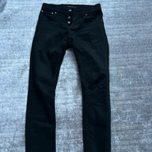 Svarta jeans från Nudie Jeans - Säljer ett par svarta grim Tim jeans från Nudie Jeans i storlek 29/30. De är i bra skick och har en klassisk design med knappar framtill. Perfekta för både vardag och fest! Skick 9/10, inga returer.