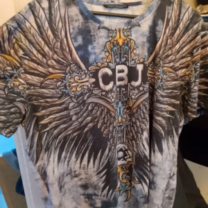 Grå t-shirt med vingar och CBJ-motiv - Säljer en cool grå t-shirt från Cipo and Baxx med ett stort CBJ-motiv och detaljerade vingar på både fram- och baksidan. T-shirten är kortärmad och har en normal passform. Perfekt för en avslappnad stil eller för att sticka ut på festen!