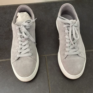 Grå sneakers i mocka - Säljer ett par snygga grå sneakers i mocka. De har en klassisk design med snörning och en vit sula som ger en fräsch look. Perfekta för vardagsbruk och passar till nästan alla outfits. Skorna är i bra skick och har knappt använts. Köp dem nu och ge din skogarderob en stilren uppdatering!