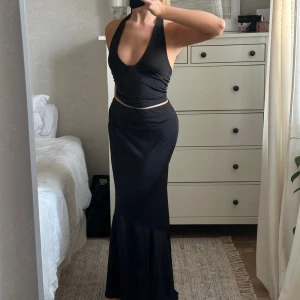 Set - Freja Wewer x NA-KD - Ett svart set, topp och kjol, från Freja Wewer x NA-KD. Toppen/linnet har en halterneckdesign. Kjolen har en resårmidja, en asymmetrisk design och har samma  skinande, stretchiga tyg som toppen. Säljes endast tillsammans. Nyskick! Använt en gång.