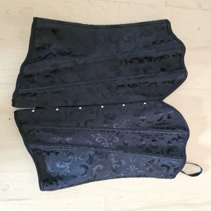 Svart korsett med mönster - Säljer en snygg svart korsett med ett elegant mönster. Den har knappar framtill och är perfekt för fest eller andra speciella tillfällen. Passformen är tight och den ger en fin siluett. Materialet känns lyxigt och hållbart.