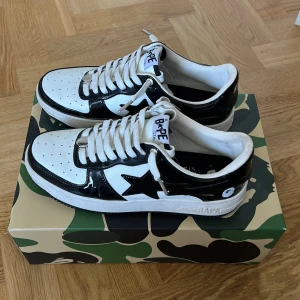 Svartvita sneakers från BAPE - Säljer ett par snygga svartvita sneakers från BAPE. Skorna är i nyskick och har en cool design med en stjärna på sidan. De är gjorda av syntetmaterial och har snörning framtill. Priset kan diskuteras! Vid snabb affär 850kr