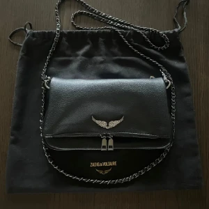 Svart axelväska från Zadig & Voltaire - Snygg svart axelväska i skinn liknande Zadig & Voltaire med kedjerem och dekorativt vingmotiv. Perfekt för att ge en edgy touch till din outfit. LÄGGER UT FÖR FRIFRAKT 