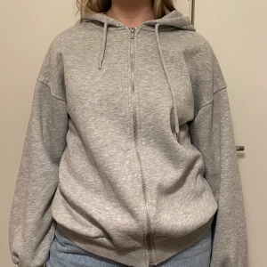 Grå hoodie från Gina Tricot - Mysig grå hoodie från Gina Tricot med dragkedja och justerbar huva utan fickor. Perfekt för en avslappnad stil. Den har en normal passform och är tillverkad i mjukt material för extra komfort.